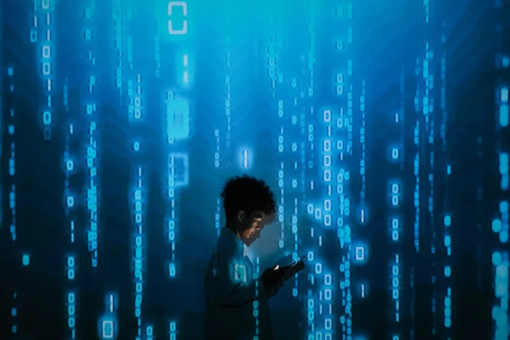 person-with-smartphone-standing-in-projection-of-zeros-and-ones Silhouette of a person using a smartphone surrounded by digital binary code projections.